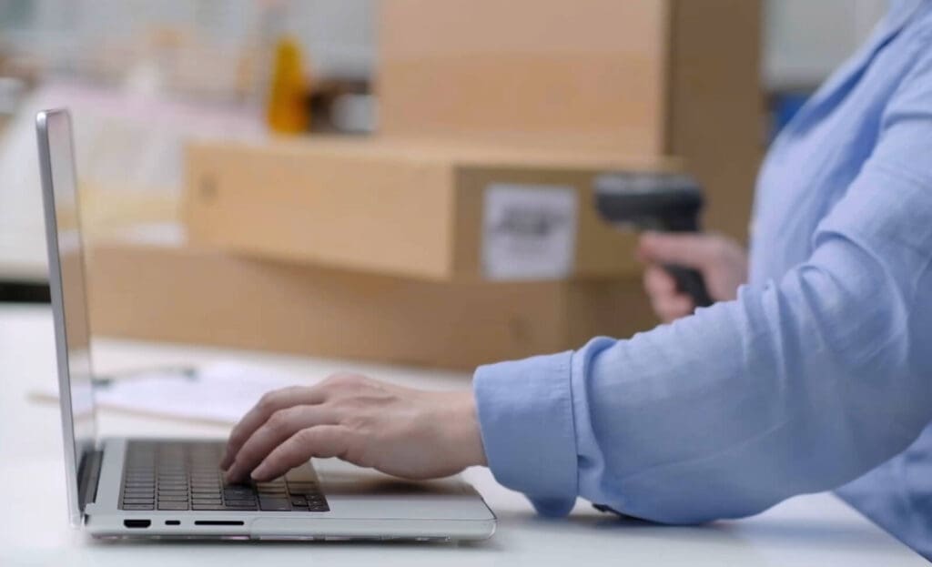 A person with a hand on a laptop and the other hand scanning a box with a scanner.